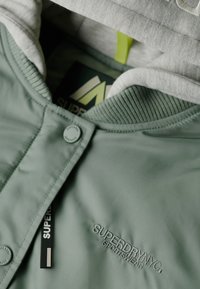 Grøn jakke med en glat finish, udstyret med hætte i grå, ribbet krave, knaplukning og broderet logo "SUPERDRY".