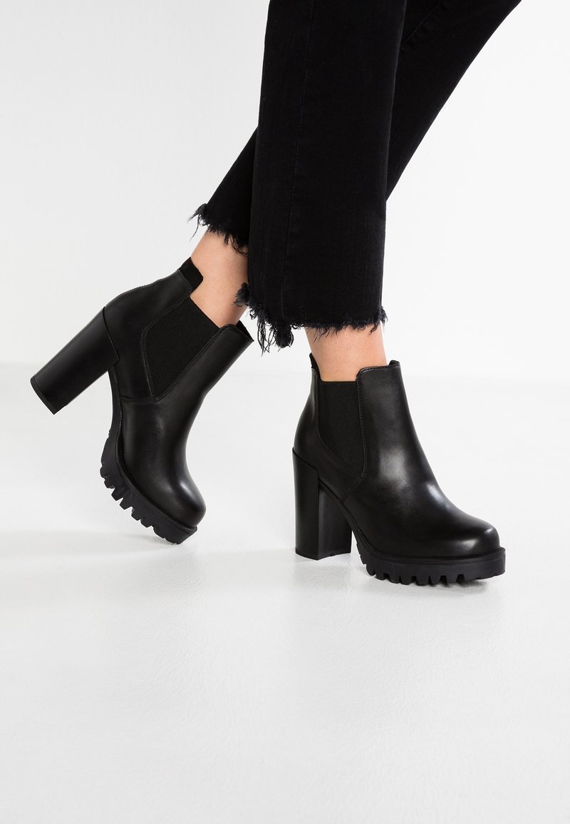 Bottines noires en cuir avec un talon carré haut, des panneaux latéraux élastiques et des semelles en caoutchouc texturé, présentant une finition lisse et élégante.