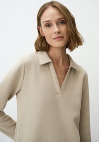 Pull en tricot beige avec un col et une encolure en V. Dispose de manches longues et d'un tissu doux et texturé. Design simple et minimal.