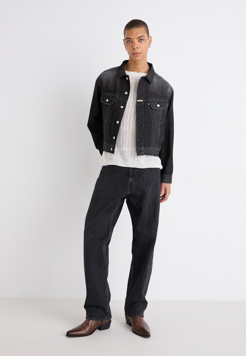 Schwarze Jeansjacke mit zwei Fronttaschen, kombiniert mit einem weißen strukturierten Pullover und schwarzen Jeans. Modell trägt braune spitze Stiefel.