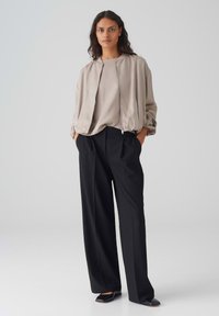 Lichtbeige bomberjack met ritssluiting, losse pasvorm en elastische manchetten, gecombineerd met high-waisted zwarte wijde broek en platte schoenen.