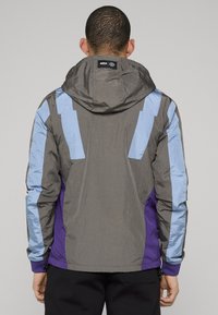 Vattentät jacka med huva, med grå kropp, blå och lila accentpaneler samt texturerat tyg. Justerbara muddar och fåll.