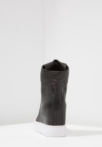 Černé kožené vysoké tenisky s bílou podrážkou, zobrazené zezadu na bílé ploše na pozadí v rohu světle šedé barvy.