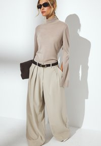 Beige Rollkragenoberteil mit langen Ärmeln, kombiniert mit weiten, hellen Hosen und einer schwarzen geflochtenen Clutch. Die Accessoires umfassen einen schwarzen Gürtel.