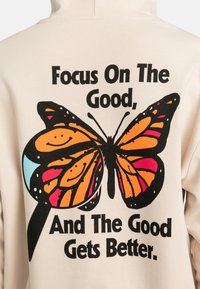 Sweatshirt crème avec un grand graphique de papillon noir et orange, portant le texte "Concentrez-vous sur le positif, et le positif s'améliore" imprimé en lettres grasses.