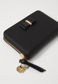 Black leather wallet with gold zipper, bow accent on the front, and gold hardware detail, featuring a charm attached to the zipper pull.