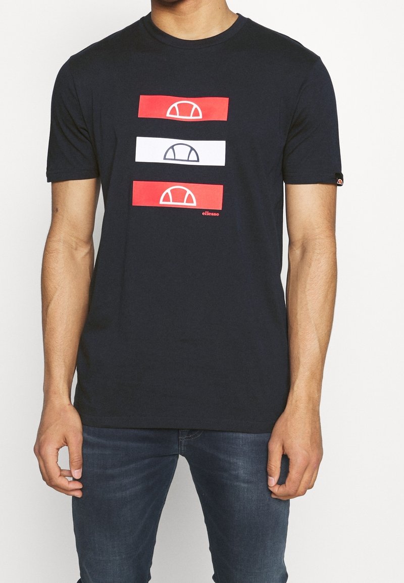 Homme portant un t-shirt noir à manches courtes avec trois bandes horizontales en rouge, blanc, rouge, chacune affichant un logo semi-circulaire, associé à un jean bleu.