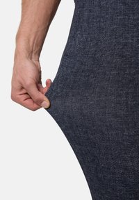 Hände, die den Stoff einer dunkelblauen, strukturierten Hose gegen einen schlichten weißen Hintergrund kneifen und dehnen.