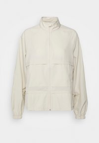 Ljusbeige zip-jacka i mjukt tyg. Har en stående krage, två bröstfickor, elastiska ärmslut och justerbar dragsko i midjan.