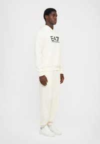 Felpa crema con logo nero, abbinata a pantaloni joggers crema coordinati. L'outfit presenta una vestibilità rilassata, polsini a coste e scarpe bianche.