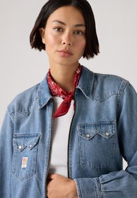 Lyseblå denimjakke med to brystlommer, sølvfargede knappeakenter og glidelås foran. Rød bandana knyttet rundt halsen. Hvit ribbet skjorte under.