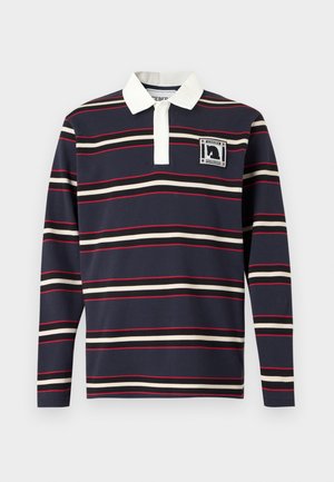 Mann trägt eine kastanienbraune Kappe, schwarzes gestreiftes Rugbyshirt mit weißem Kragen, graue weite Hose und schwarze Schuhe, steht vor einem einfarbigen Hintergrund.