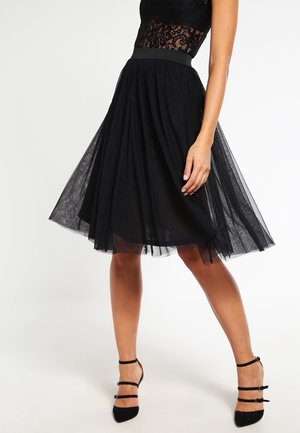 Mujer con falda negra de tul hasta la rodilla, top de encaje y tacones negros con correas, de pie con una pierna doblada y la mano apoyada en la falda.