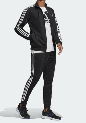 Personne portant un survêtement Adidas noir avec trois bandes blanches sur les manches et les pantalons, un t-shirt blanc avec le logo Adidas, et des baskets noires.
