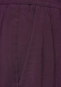 Tessuto viola scuro che mette in mostra una leggera lucentezza, con dettagli in filo e una tasca laterale. Texture liscia con variazioni sottili.