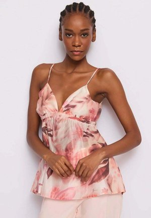 Donna con capelli intrecciati che indossa un top senza maniche rosa a fiori con spalline sottili, in piedi davanti a uno sfondo chiaro e semplice.