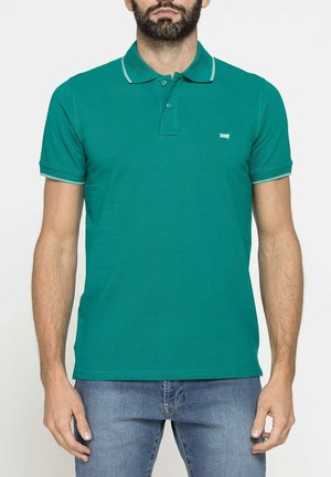 Uomo con barba che indossa una polo tinta unita color verde acqua a maniche corte e jeans blu, in piedi davanti a uno sfondo bianco.