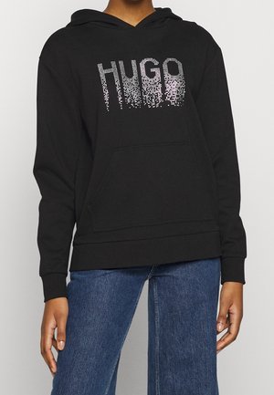 Czarna bluza z kapturem z przednią kieszenią i napisem "HUGO" w blednących białych kropkach, noszona z niebieskimi dżinsami.