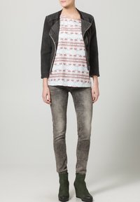 Veste noire texturée, blouse blanche avec motif flamant rose, jeans skinny gris et bottes compensées vertes. Design minimaliste, ensemble décontracté.