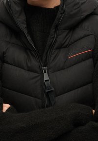 Svart quiltad puffer väst med huva, försedd med en hel dragkedja, Superdry-logotyp i reflekterande grått och rött, ihop med en svart stickad tröja.