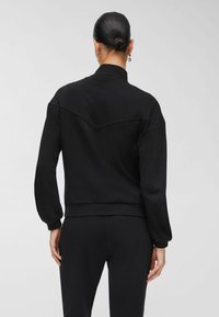 Sweat-shirt noir avec un col montant et des manches longues. Présente un détail d'empiècement incurvé au dos, fabriqué en tissu doux et flexible.