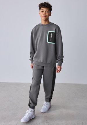 Adolescente con chándal gris con detalle en el bolsillo en negro y verde menta, zapatillas blancas, de pie sobre suelo gris con fondo gris claro.