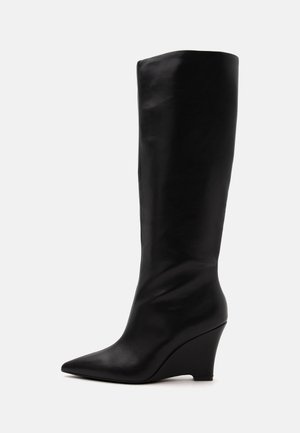 Botte en cuir noire montant au genou avec bout pointu et talon compensé, vue de côté sur un fond clair uni.