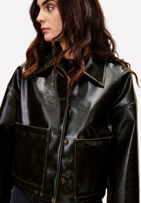 Vrouw met golvend bruin haar, gekleed in een glanzende donkergroene leren jas met grote voordo pockets en bruine knopen, kijkend naar links.