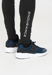 Zwarte leggings met het woord "ENDURANCE" in het wit gedrukt, gecombineerd met blauwe en zwarte sport schoenen met een mesh bovenkant en een witte zool.
