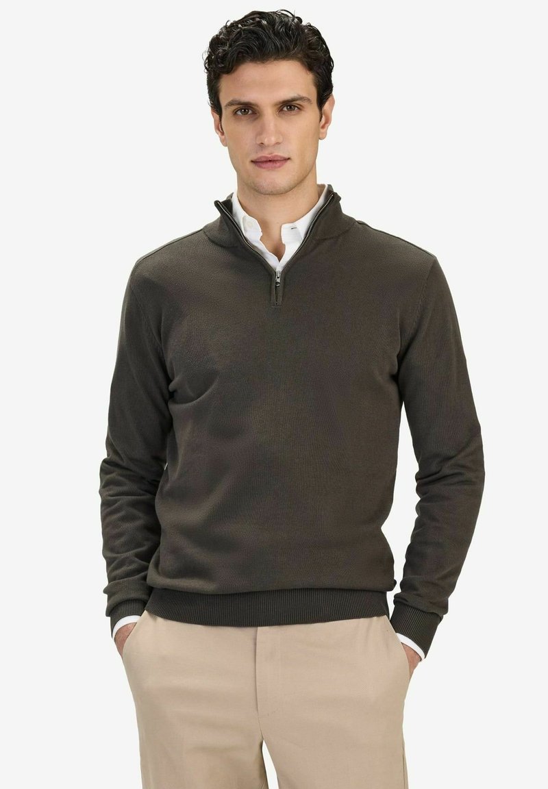 Homme portant un pull à fermeture éclair quart kaki foncé par-dessus une chemise blanche et un pantalon beige, debout les mains dans les poches devant un fond uni.