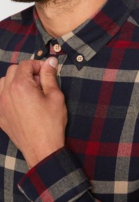 Koszula flanelowa z wzorem w kratę w kolorach granatowym, czerwonym i beżowym. Kołnierzyk zapinany na guziki, zapięcie na dwa guziki z przodu oraz fakturowana tkanina.