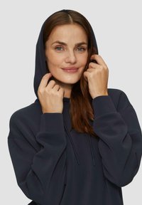 Donkergrijze hoodie met lange mouwen, koord en zachte textuur, gedragen door een persoon die de capuchon aanpast.