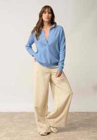 Jasnoniebieski sweter dzianinowy z głębokim dekoltem w kształcie litery V, zestawiony z szerokimi, beżowymi spodniami z kordurowej tkaniny, zdobionymi pionowymi pasami i luźnym fasonem.