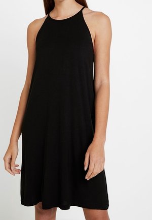 Femme portant une robe noire sans manches, longueur genou, avec un col montant, debout devant un fond blanc uni.