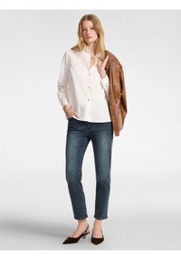 Camicia bianca con colletto, abbinata a jeans blu scuro. L'outfit include una giacca di pelle marrone appoggiata su un braccio e scarpe marroni.