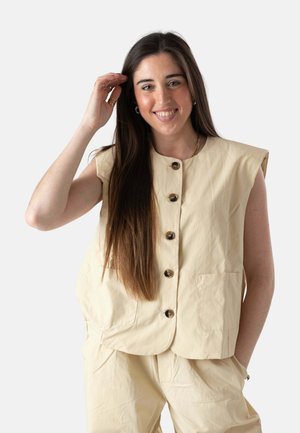 Mujer sonriente con cabello largo y castaño que lleva un chaleco beige sin mangas abotonado y pantalones a juego, con una mano tocándose el cabello, sobre un fondo liso.