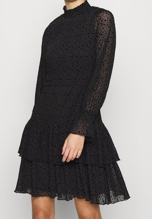 Czarna przezroczysta sukienka z długim rękawem z aksamitnymi groszkami, wysokim kołnierzem, marszczonymi mankietami i warstwową spódnicą z falbanami, noszona przez osobę.