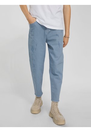 Osoba oblečená v světle modrých roztrhaných kalhotách, béžových šněrovacích botách a bílém tričku, stojící s jednou rukou v kapse.