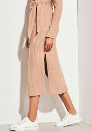 Robe midi en tricot de couleur rose clair en jersey, avec une taille cintrée et des fentes sur les côtés. Assortie à des baskets blanches. Texture lisse, design ajusté.