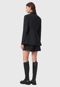 Zwarte op maat gemaakte blazer met peplumontwerp, gecombineerd met zwarte shorts en kniehoge leren laarzen. Glad stof en slanke silhouet.