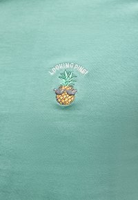 Μικρός κεντημένος ανανάς που φοράει γυαλιά ηλίου με το κείμενο «LOOKING PINE1» σε πράσινο υφασμάτινο φόντο.