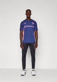 Man die naar voren kijkt en een blauw-roze sportshirt, zwarte sportbroek en witte sneakers draagt tegen een effen achtergrond.