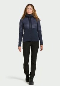 Námornícky modrá bunda so zipsom a kapucňou; obsahuje prešívané horné časti a hladký dolný panel. Skombinovaná s čiernymi nohavicami a čižmami.