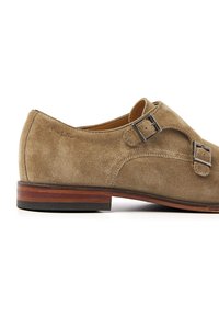 Tan suede shoe with dual buckles, rounded toe, and a wooden heel. Features a smooth texture and minimal stitching detail.