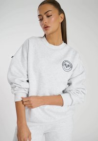 Lichtgrijze sweatshirt met een ronde halslijn, ribbels aan de mouwen en een geborduurd logo van de tennisclub op de borst. Zachte, gestructureerde stof.