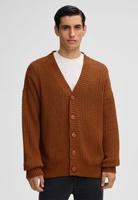 Cardigan lavorato a maglia marrone con scollo a V, polsini a coste e cinque grandi bottoni. Presenta motivi testurizzati su tutta la stoffa.