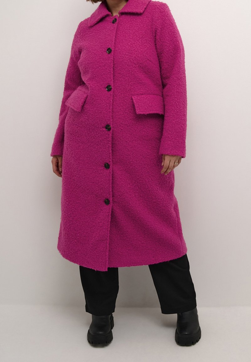 Kobieta ubrana w długi, teksturowany jasnoróżowy płaszcz na guziki z klapkowymi kieszeniami, czarne spodnie i czarne masywne botki, stojąca na jednolitym tle.