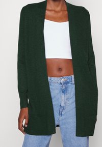 Ciemnozielony kardigan o prążkowanej teksturze, z otwartym przodem, długimi rękawami i dwiema przednimi kieszeniami. Nosi się go na białym topie crop i niebieskich dżinsach.