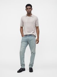 Un homme se tient debout, une main dans la poche, vêtu d’un t-shirt en maille beige clair à manches courtes, d’un jean bleu clair et de chaussures noires sur un fond uni.