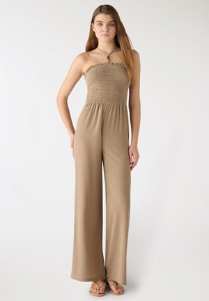 Donna in piedi che indossa una tuta senza spalline color beige con pantaloni a gamba larga e sandali su uno sfondo bianco semplice.